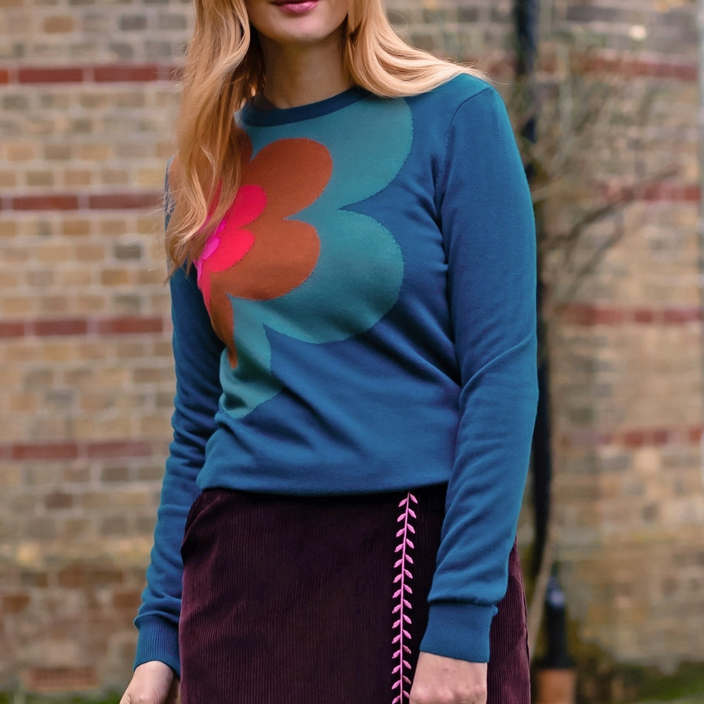 A woman wearing a teal long-sleeve sweater with a large retro floral pattern in orange and teal, paired with a dark purple skirt, standing outdoors in front of a brick wall.
