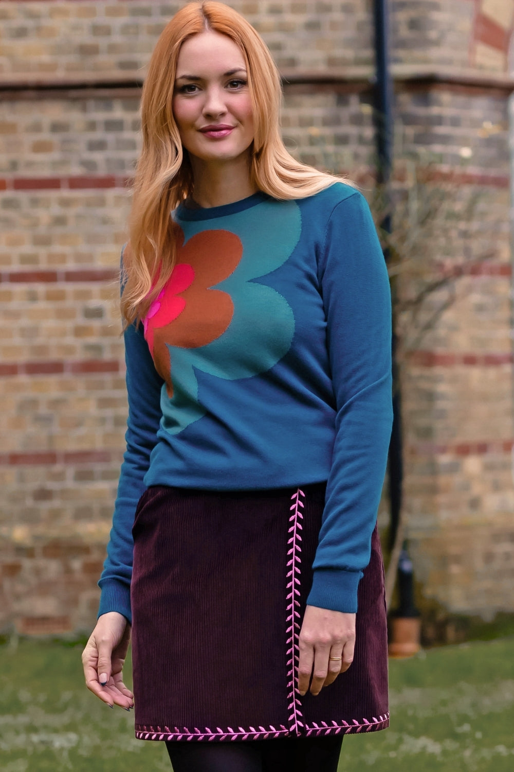 A woman wearing a teal long-sleeve sweater with a large retro floral pattern in orange and teal, paired with a dark purple skirt, standing outdoors in front of a brick wall.