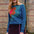 A woman wearing a teal long-sleeve sweater with a large retro floral pattern in orange and teal, paired with a dark purple skirt, standing outdoors in front of a brick wall.