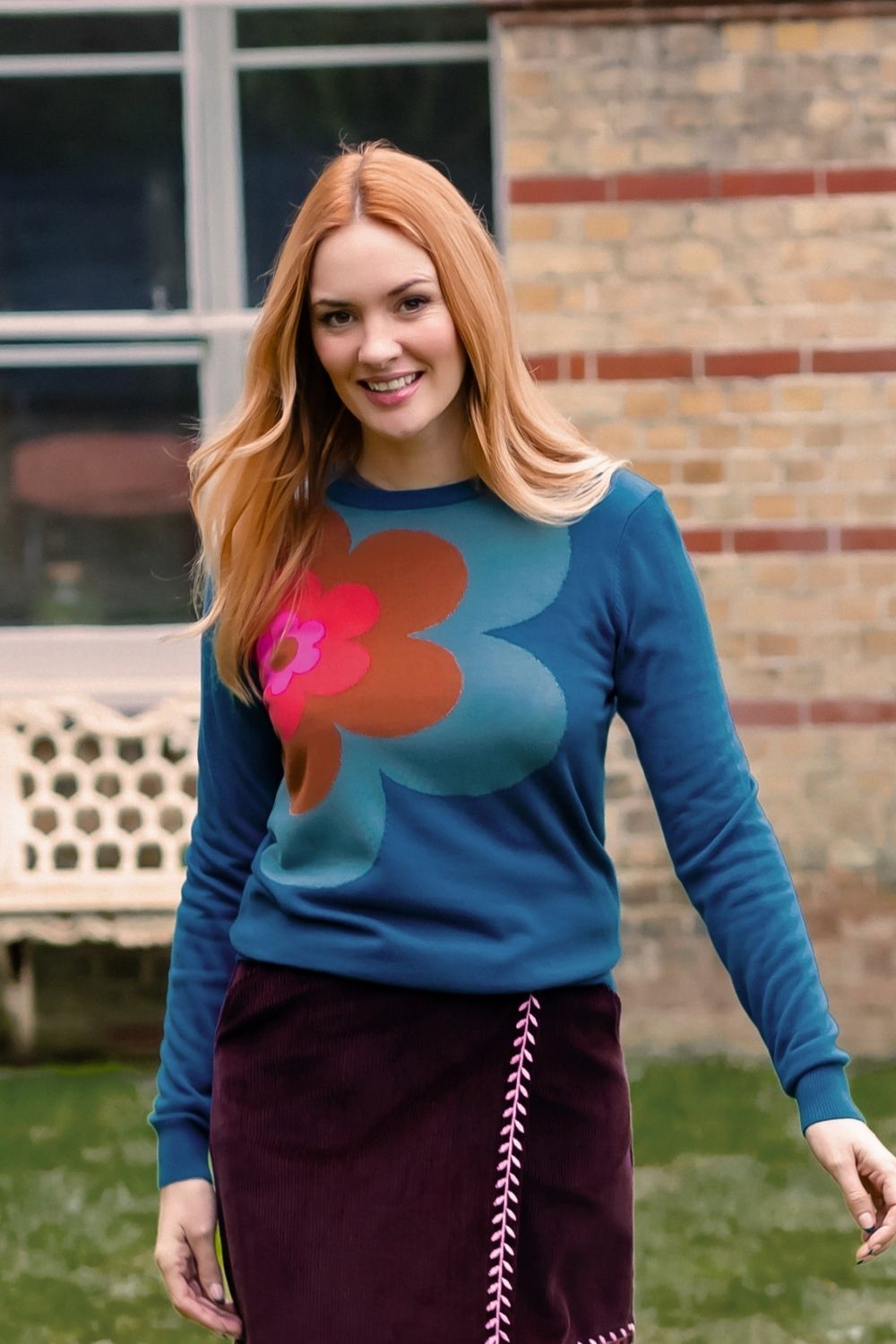 A woman with long red hair wearing a teal long-sleeve crewneck sweater with a large retro flower graphic in pink, orange, and teal, paired with a dark burgundy skirt. She is standing outdoors in front of a brick building and green grass.