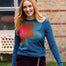 A woman with long red hair wearing a teal long-sleeve crewneck sweater with a large retro flower graphic in pink, orange, and teal, paired with a dark burgundy skirt. She is standing outdoors in front of a brick building and green grass.