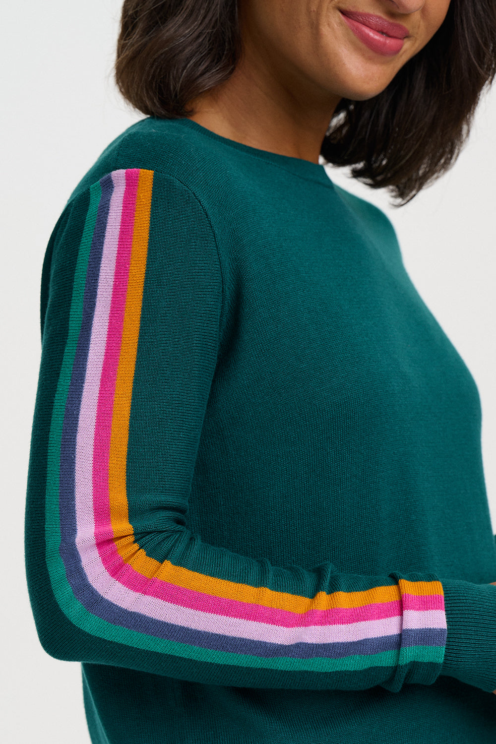 A close-up of a woman wearing a dark teal green knit sweater with a vibrant, multicolored striped ribbed sleeve detail on the upper arm and forearm.