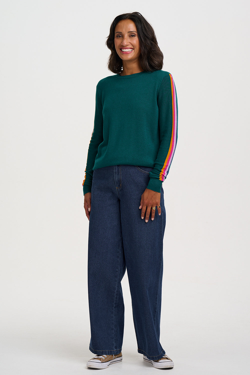 A woman modeling a dark green long-sleeve knit sweater with a vibrant rainbow stripe down one sleeve, paired with dark blue wide-leg jeans and white sneakers, against a plain light gray studio background.