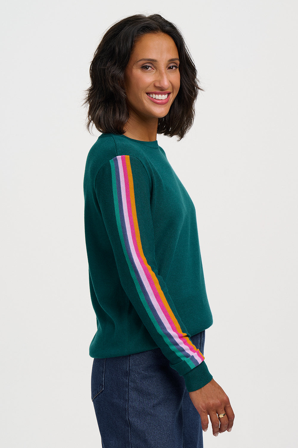 A woman wearing a dark green crewneck sweatshirt with a vibrant rainbow stripe detail down the sleeves, paired with dark blue jeans, photographed against a solid light gray background.