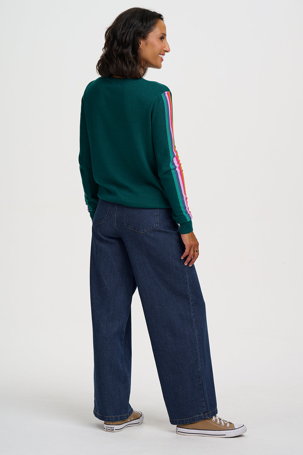 A woman shown from the back wearing a dark green long-sleeve sweater with a colorful striped detail on the sleeve, dark blue wide-leg jeans, and tan sneakers, against a plain light gray studio background.