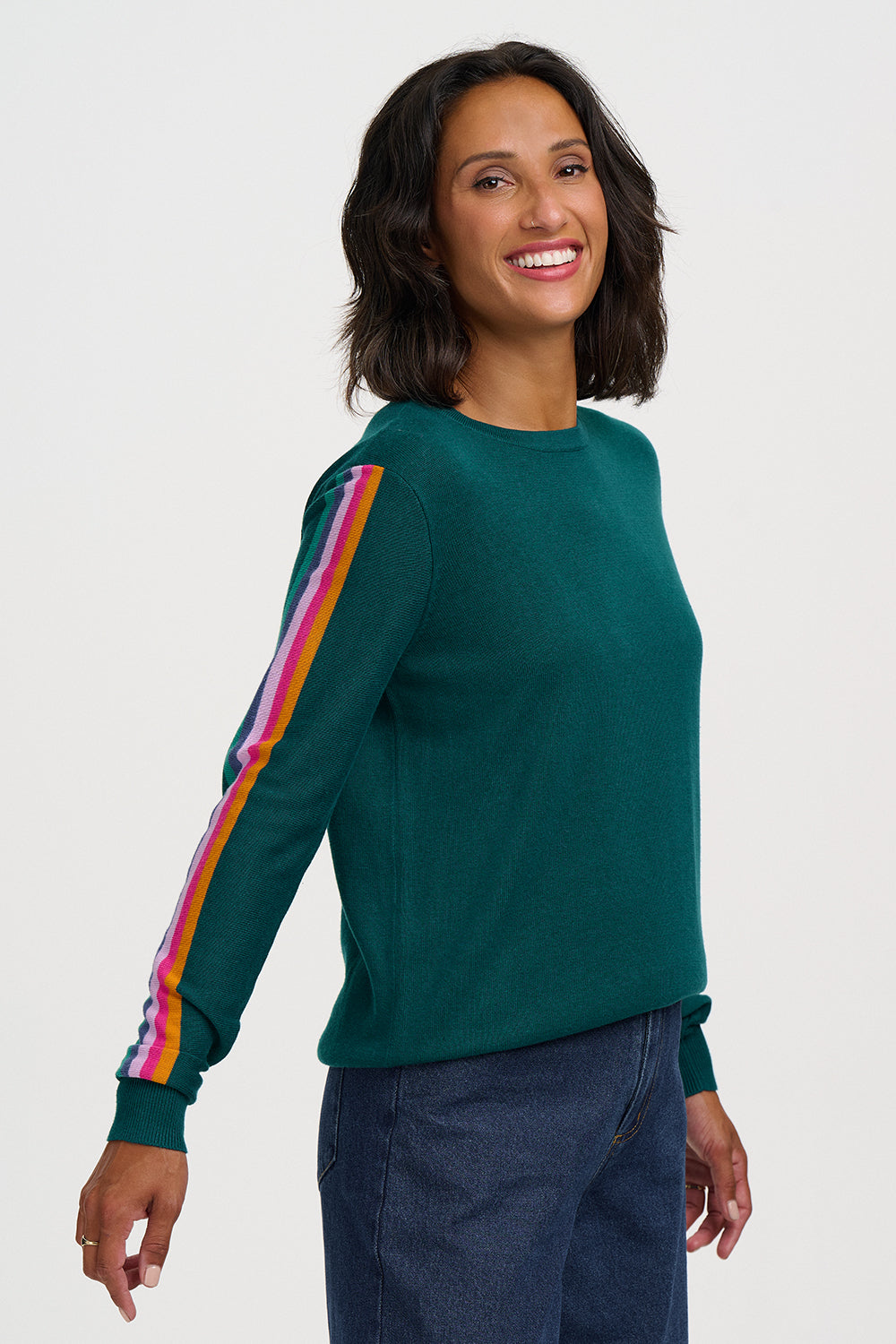 A woman wearing a dark green crewneck sweater with a vibrant, multicolored striped accent down one sleeve, paired with dark blue jeans, against a plain light gray studio background.