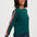 A woman wearing a dark green crewneck sweater with a vibrant, multicolored striped accent down one sleeve, paired with dark blue jeans, against a plain light gray studio background.