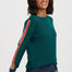 A woman wearing a dark green crewneck sweater with a vibrant, multicolored striped accent down one sleeve, paired with dark blue jeans, against a plain light gray studio background.