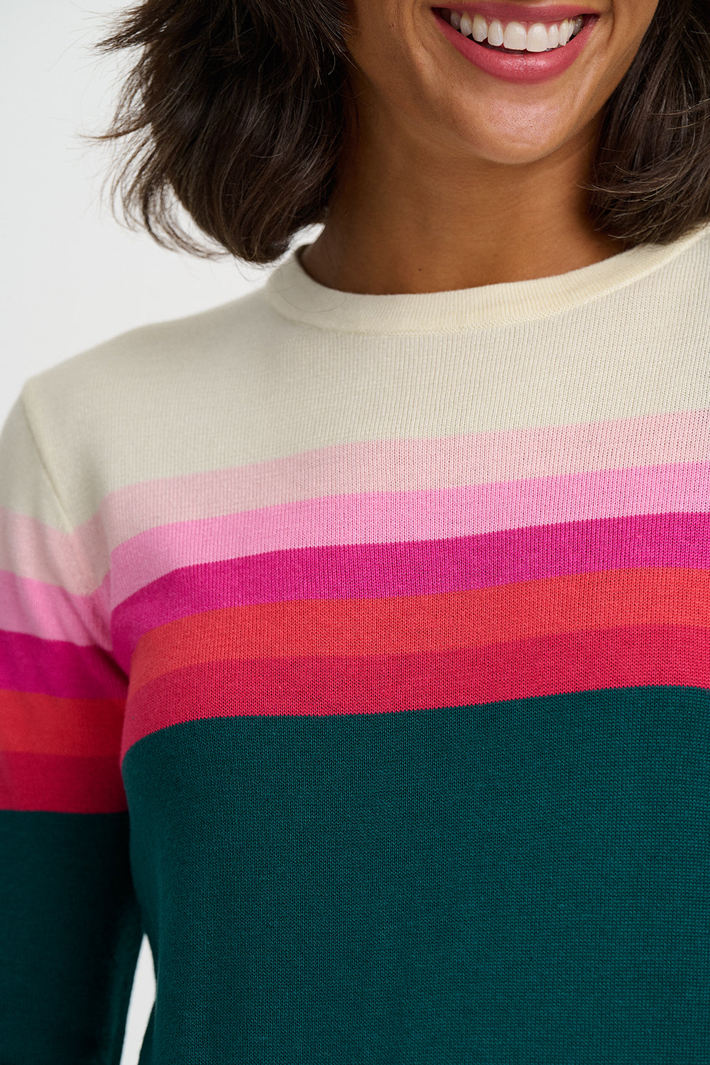 A close-up of a woman wearing a ribbed knit crewneck sweater with horizontal stripes in cream, light pink, darker pink, coral red, and dark teal green.