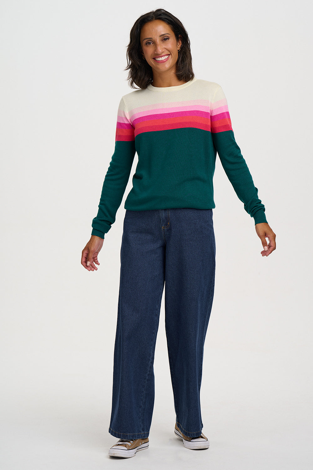 A woman modeling a long-sleeve crewneck sweater with horizontal stripes in cream, light pink, hot pink, and dark green, paired with dark blue wide-leg jeans and white sneakers, against a light gray studio background.