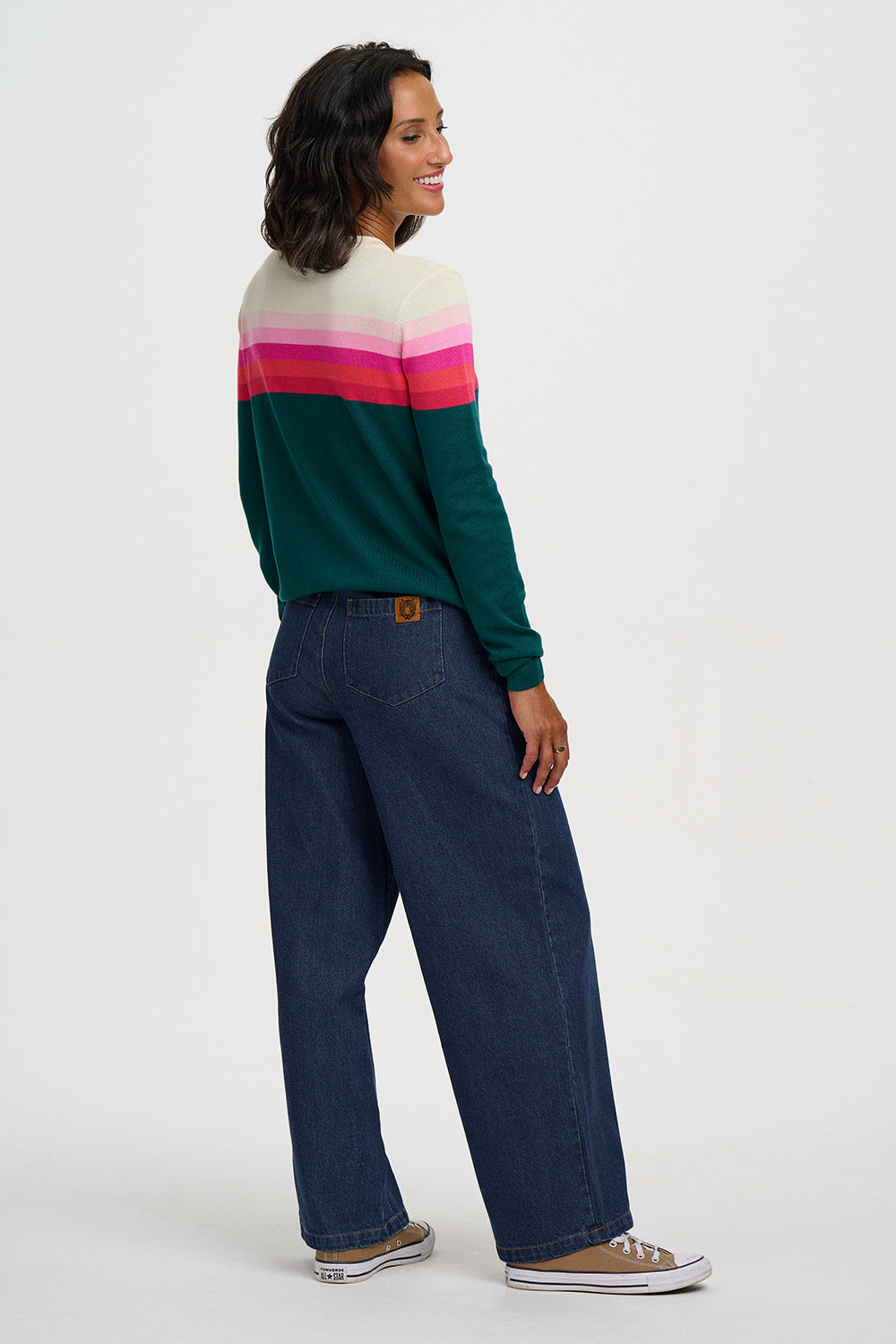 A woman is shown from the back wearing a color-blocked knit sweater with dark green, pink, and cream horizontal stripes, paired with dark blue wide-leg jeans and brown sneakers, against a plain light gray studio background.