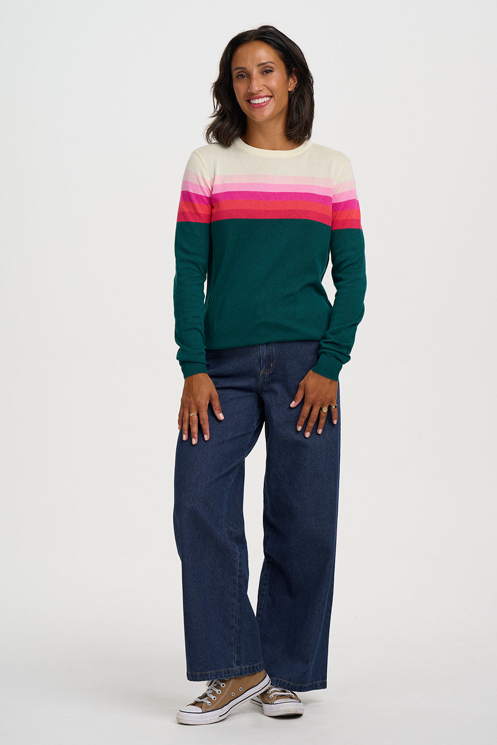 A woman modeling a long-sleeve crewneck sweater with horizontal stripes in cream, white, pink, and dark green, paired with dark blue jeans and brown sneakers, against a plain light gray studio background.