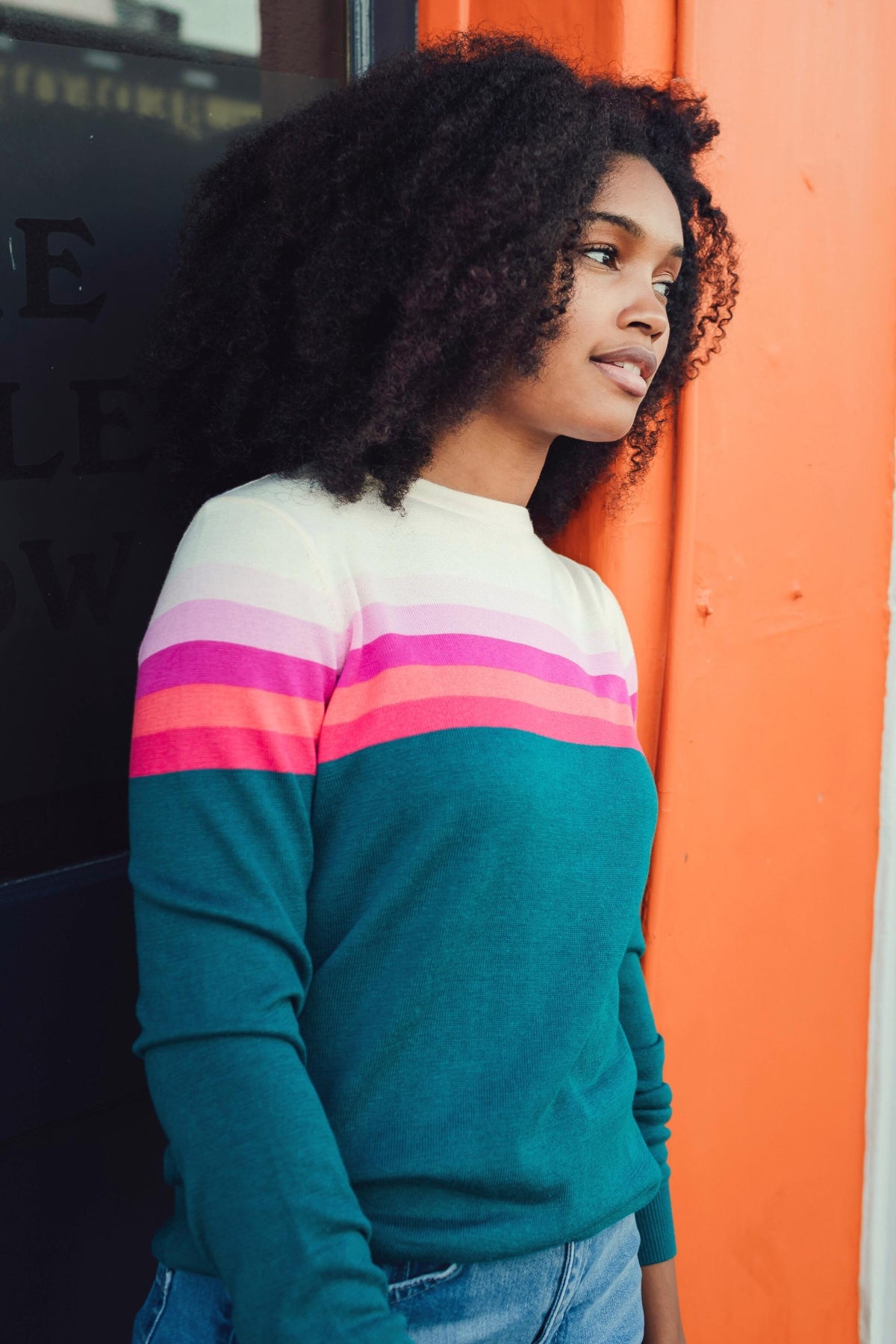A woman with curly black hair wearing a teal crewneck sweater with horizontal stripes in cream, light pink, magenta, and coral, standing next to a bright orange wall.