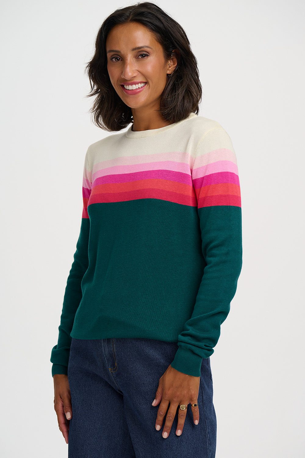 A woman wearing a color-blocked knit sweater with a cream upper chest, horizontal stripes in white, pink, and magenta across the chest and upper arms, and solid dark green sleeves and body, paired with dark blue jeans against a light gray studio background.