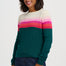 A woman wearing a color-blocked knit sweater with a cream upper chest, horizontal stripes in white, pink, and magenta across the chest and upper arms, and solid dark green sleeves and body, paired with dark blue jeans against a light gray studio background.