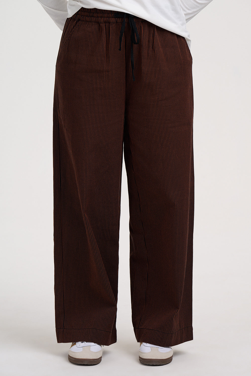 A person wearing dark brown wide-leg pleated trousers with a black drawstring waist, styled with a white top and white sneakers against a light gray studio background.