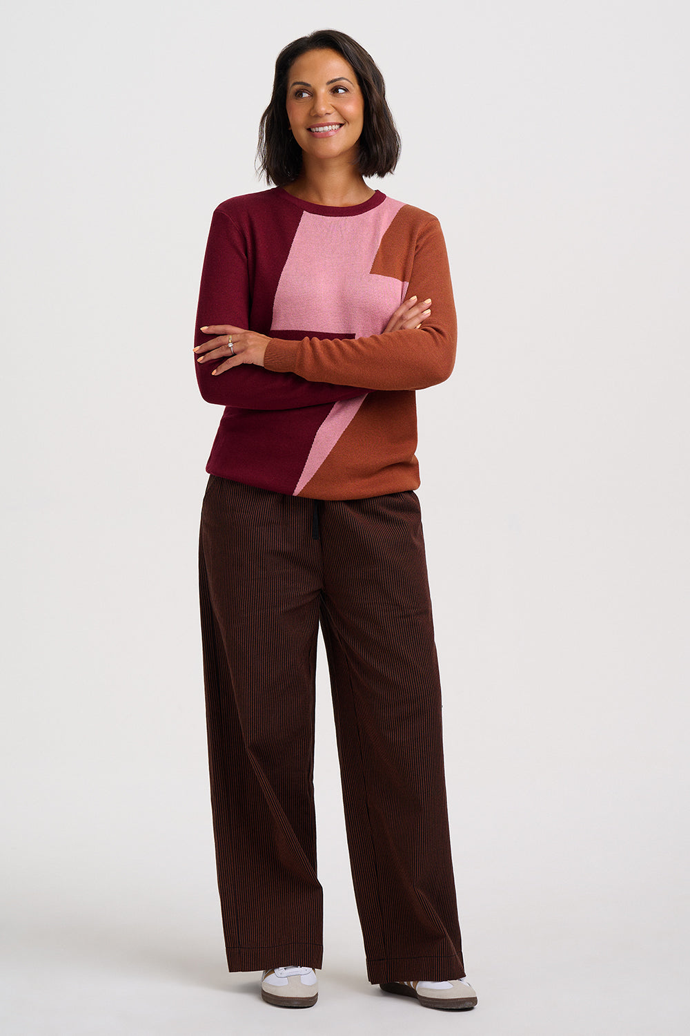 A woman modeling a color-blocked knit sweater with wide-leg brown trousers, standing against a plain light gray studio background.