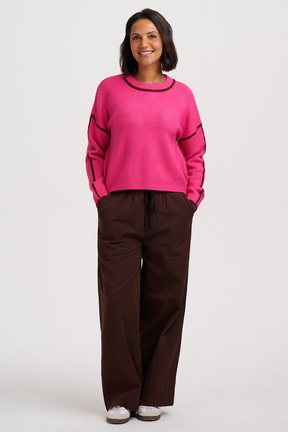 A woman modeling a bright pink crewneck sweater with black trim and dark brown wide-leg pants against a light gray studio background.