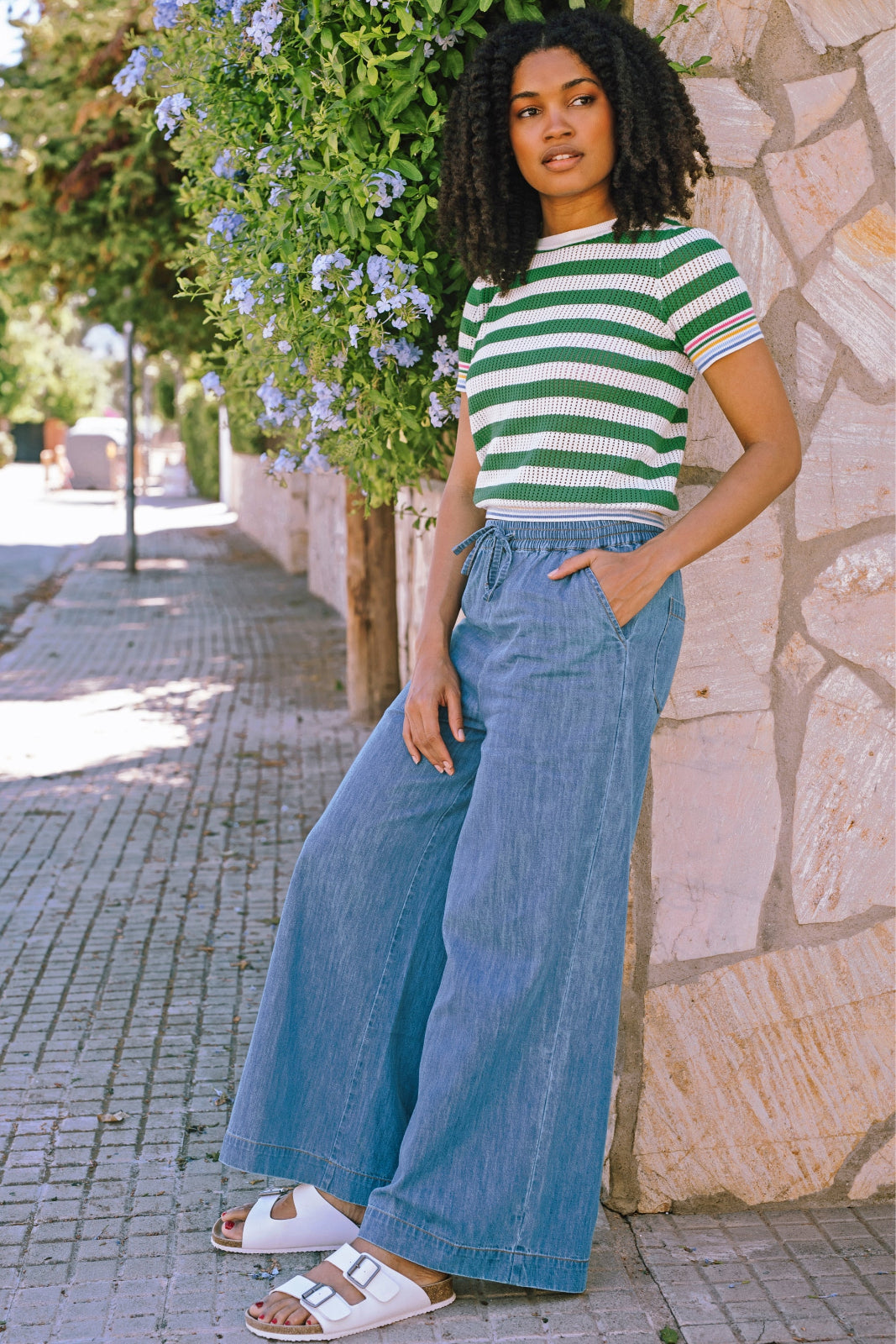 A model wearing mid blue wide leg trousers with an elasticated drawstring waist, paired with a striped top and white sandals.