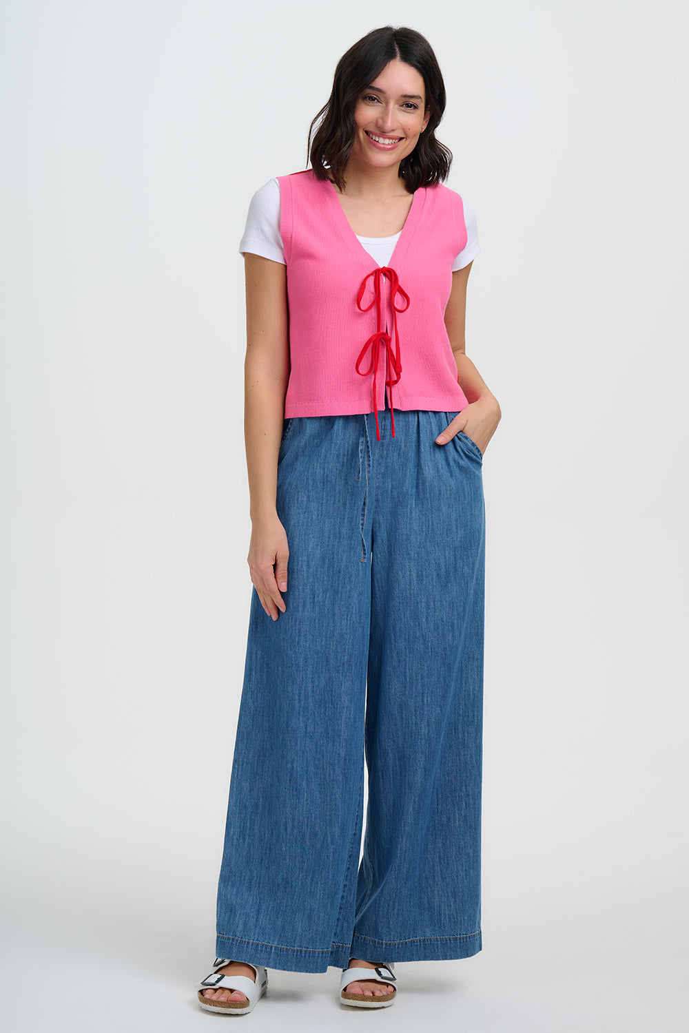 Woman wearing a pink vest over a white shirt with blue wide-leg pants on a white background