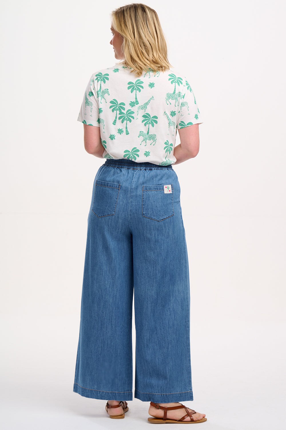 A model wearing mid blue denim wide leg trousers with an elasticated drawstring waist and back patch pockets.