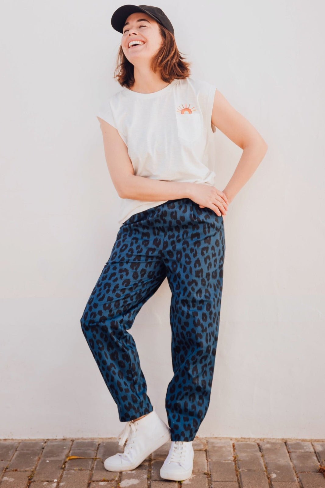 A person is standing, wearing Romney Trousers in a petrol navy color with an ikat leopard print design, paired with a white t-shirt and white sneakers.