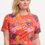 A woman wearing a red crew neck t-shirt with a painted jungle print, featuring vibrant colors and tropical motifs.