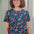 A woman wearing a teal blue crew neck t-shirt with a floral and hidden dinosaur pattern, paired with sunglasses and blue jeans.