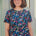 A woman wearing a teal blue crew neck t-shirt with a floral and hidden dinosaur pattern, paired with sunglasses and blue jeans.