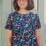A woman wearing a teal blue crew neck t-shirt with a floral and hidden dinosaur pattern, paired with sunglasses and blue jeans.