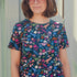 A woman wearing a teal blue crew neck t-shirt with a floral and hidden dinosaur pattern, paired with sunglasses and blue jeans.