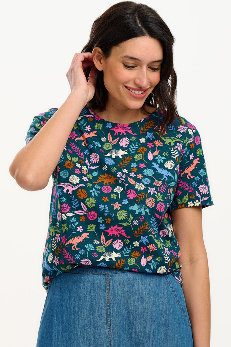 A woman smiling and wearing a teal t-shirt with a floral and dinosaur pattern, paired with a denim skirt.