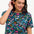 A woman smiling and wearing a teal t-shirt with a floral and dinosaur pattern, paired with a denim skirt.