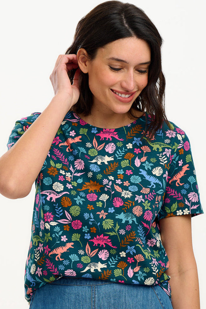 A woman smiling and wearing a teal t-shirt with a floral and dinosaur pattern, paired with a denim skirt.