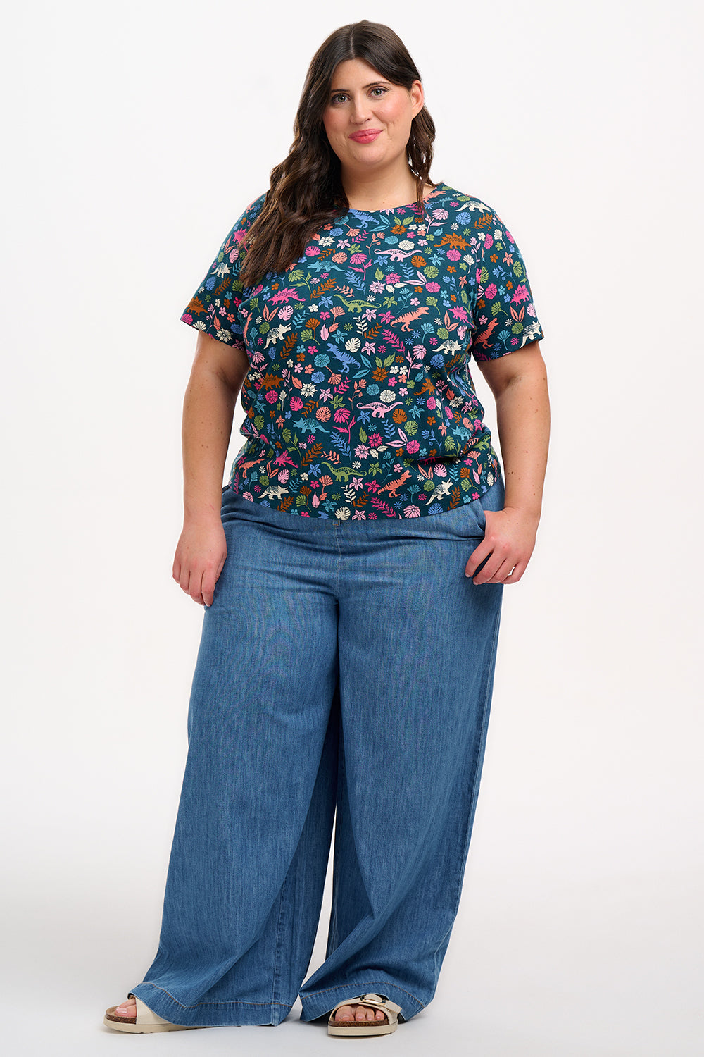 A woman smiling and wearing a teal t-shirt with a floral and dinosaur pattern, paired with a denim skirt.