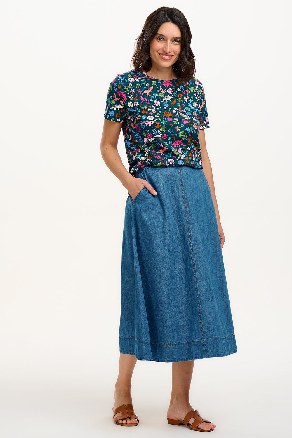 A woman smiling and wearing a teal t-shirt with a floral and dinosaur pattern, paired with a denim skirt.