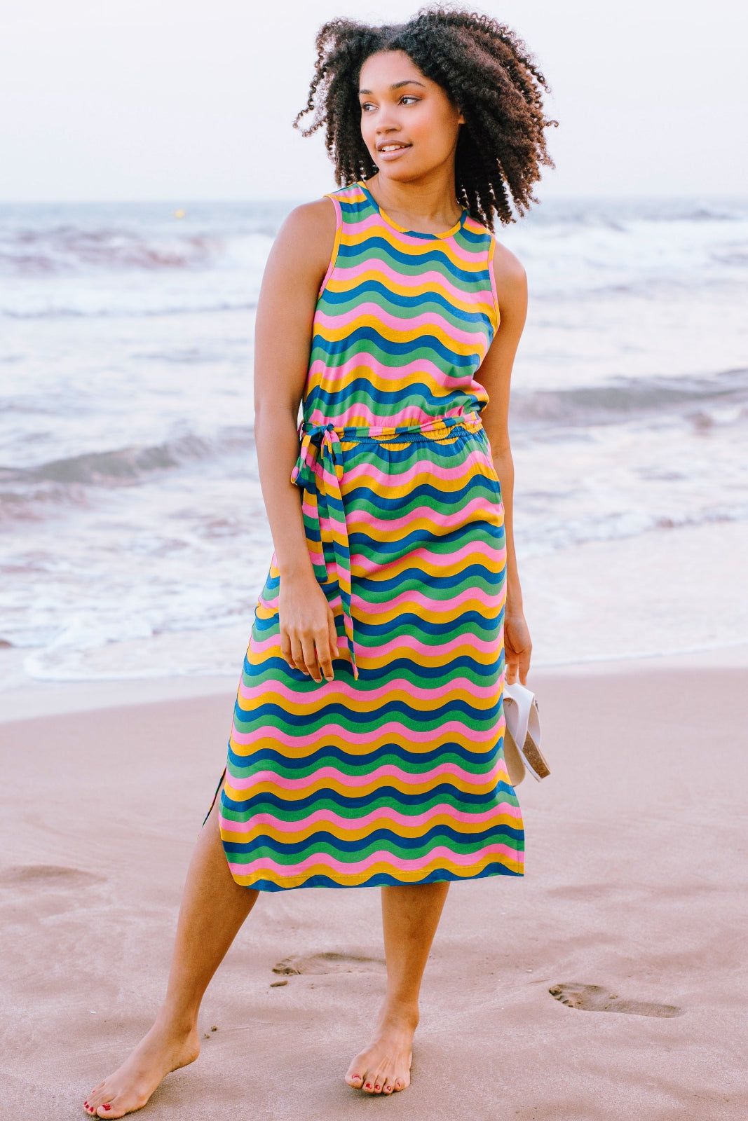 A woman standing on the beach wearing a sleeveless, midi-length, multicolored jersey dress with a pattern of rainbow waves in shades of pink, green, blue, and tan.