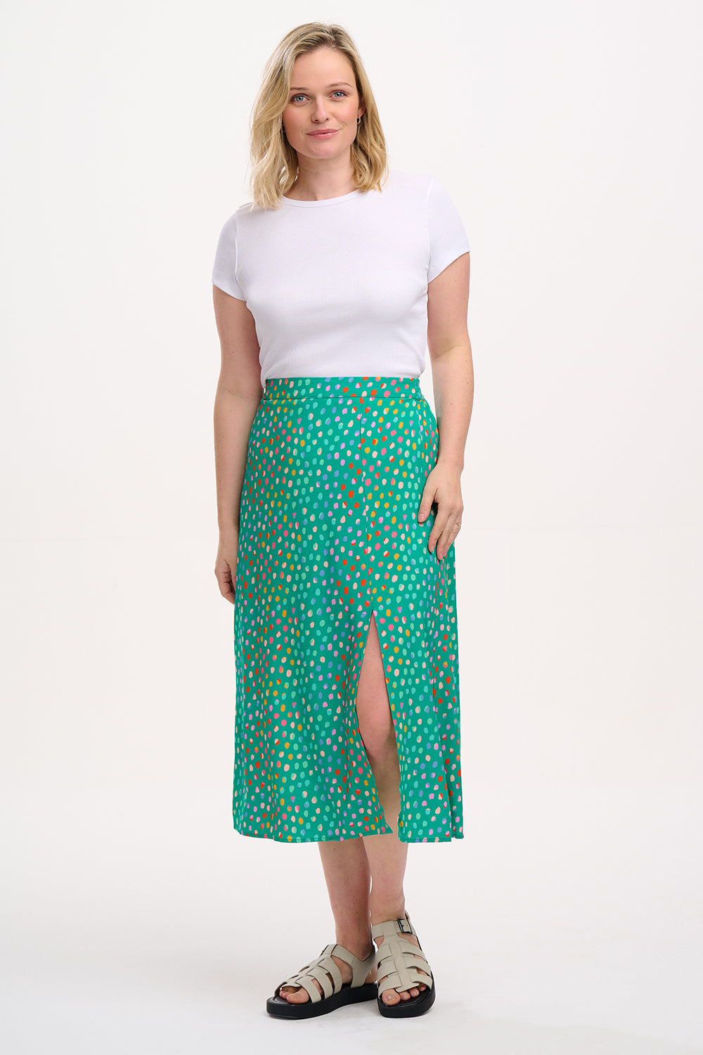 A woman standing, wearing a white top and the Rosalind Skirt in green with a rainbow pebbles print.