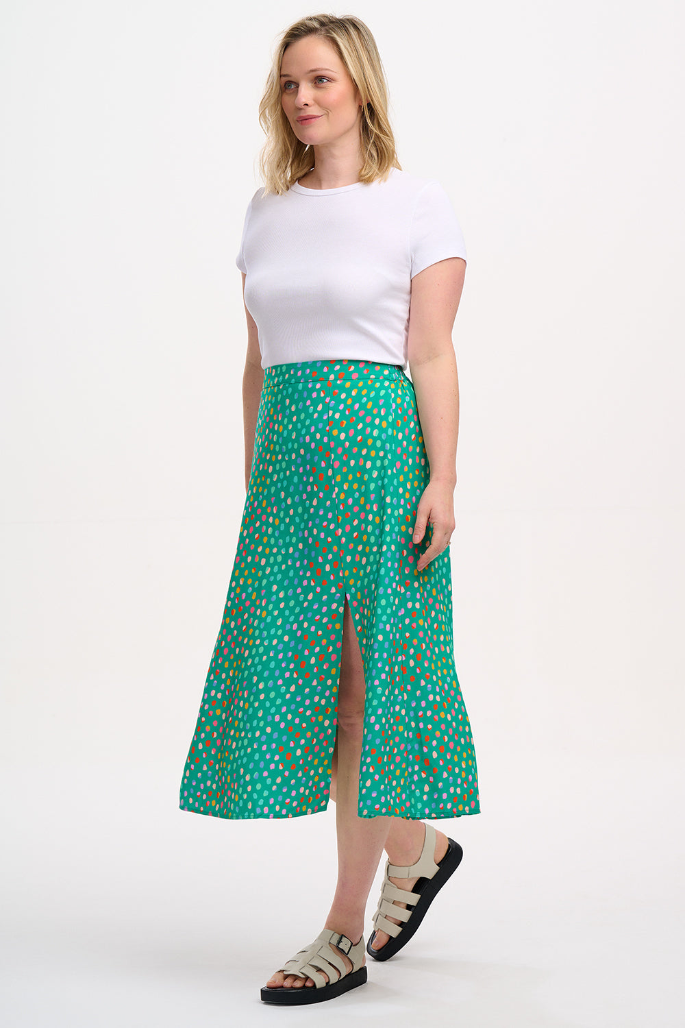 A woman standing, wearing a white top and the Rosalind Skirt in green with a rainbow pebbles print.