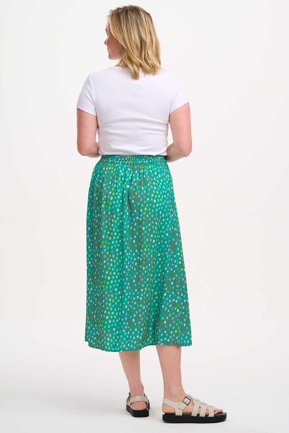 A woman standing, wearing a white top and the Rosalind Skirt in green with a rainbow pebbles print.
