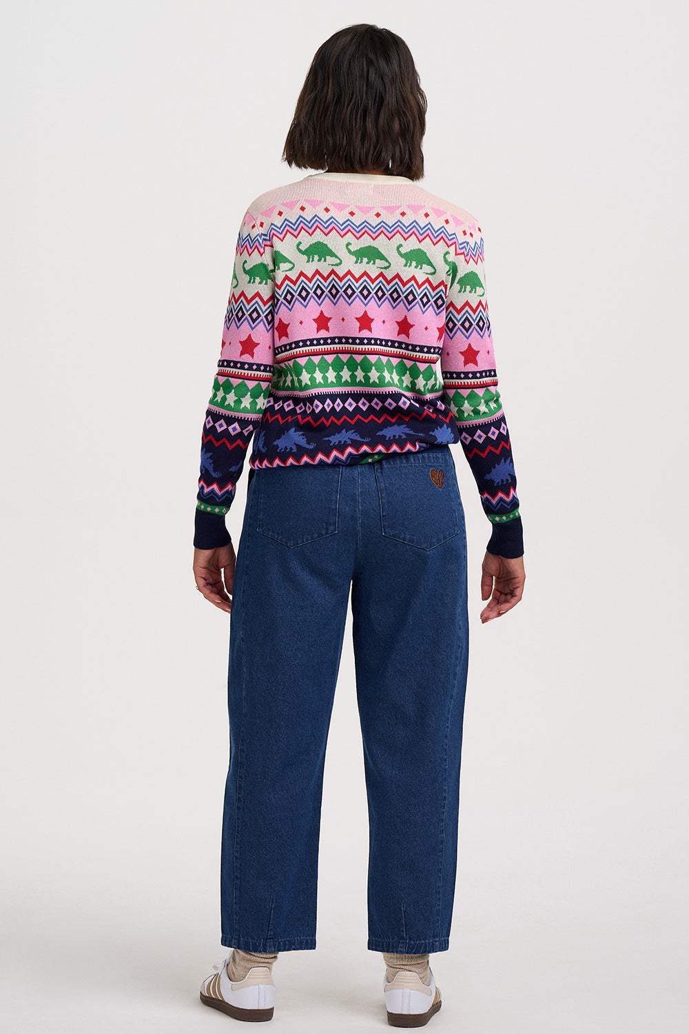 A model viewed from the back wearing a pink and multicolor Fair Isle patterned sweater with green dinosaur motifs and dark blue jeans, set against a plain white studio background.