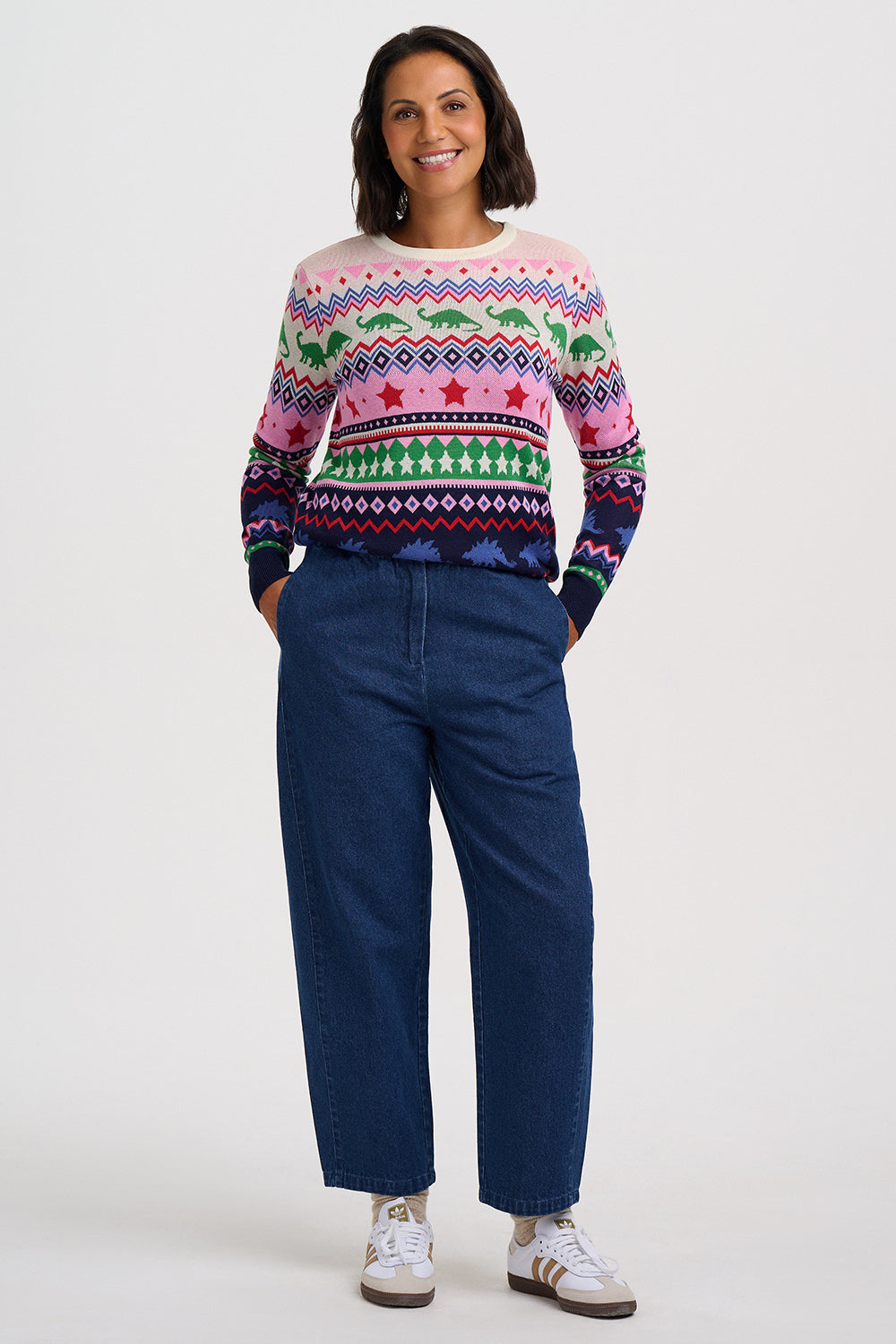 A woman modeling a long-sleeve crewneck sweater with a colorful geometric and dinosaur pattern, paired with dark blue jeans and white sneakers, against a light gray studio background.