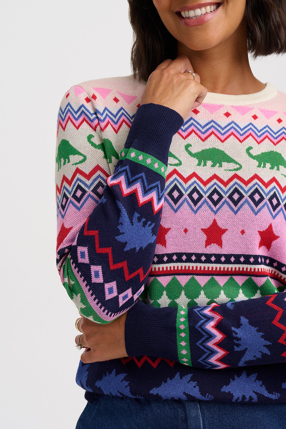 A close-up of a woman wearing a colorful Christmas sweater with dinosaur and geometric patterns in pink, navy, green, red, and white.