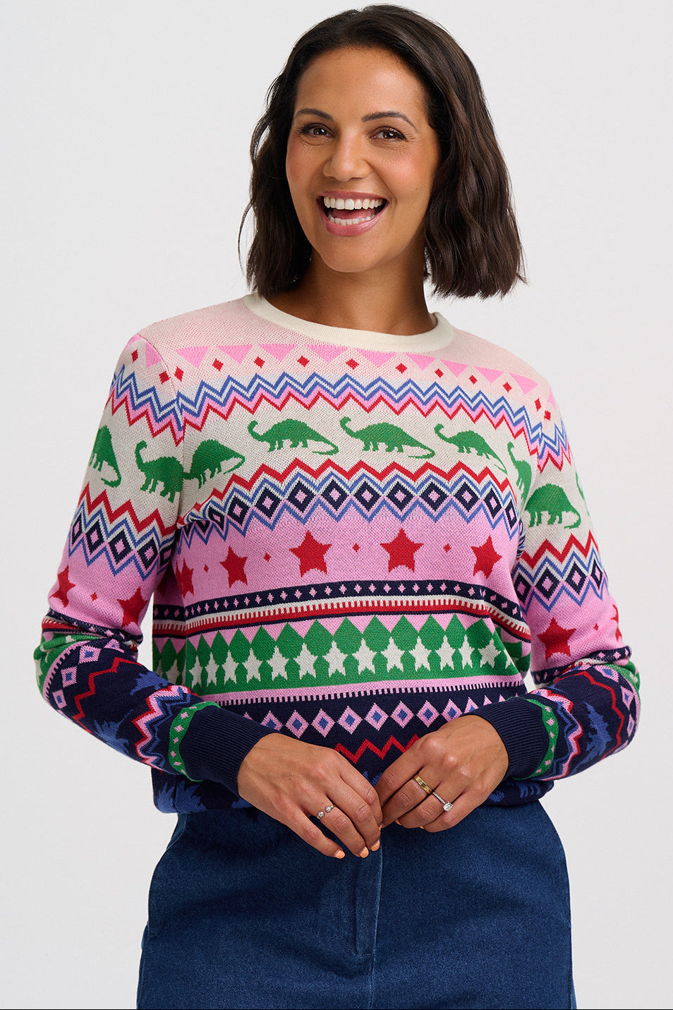 A woman smiling while wearing a long-sleeve crewneck sweater with a colorful geometric and dinosaur pattern, paired with a dark blue denim skirt, against a light gray studio background.