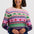 A woman smiling while wearing a long-sleeve crewneck sweater with a colorful geometric and dinosaur pattern, paired with a dark blue denim skirt, against a light gray studio background.