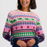 A woman smiling while wearing a long-sleeve crewneck sweater with a colorful geometric and dinosaur pattern, paired with a dark blue denim skirt, against a light gray studio background.