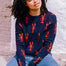 A person sitting on the ground wearing a navy crew neck jumper with a bold lobster repeat pattern in red.