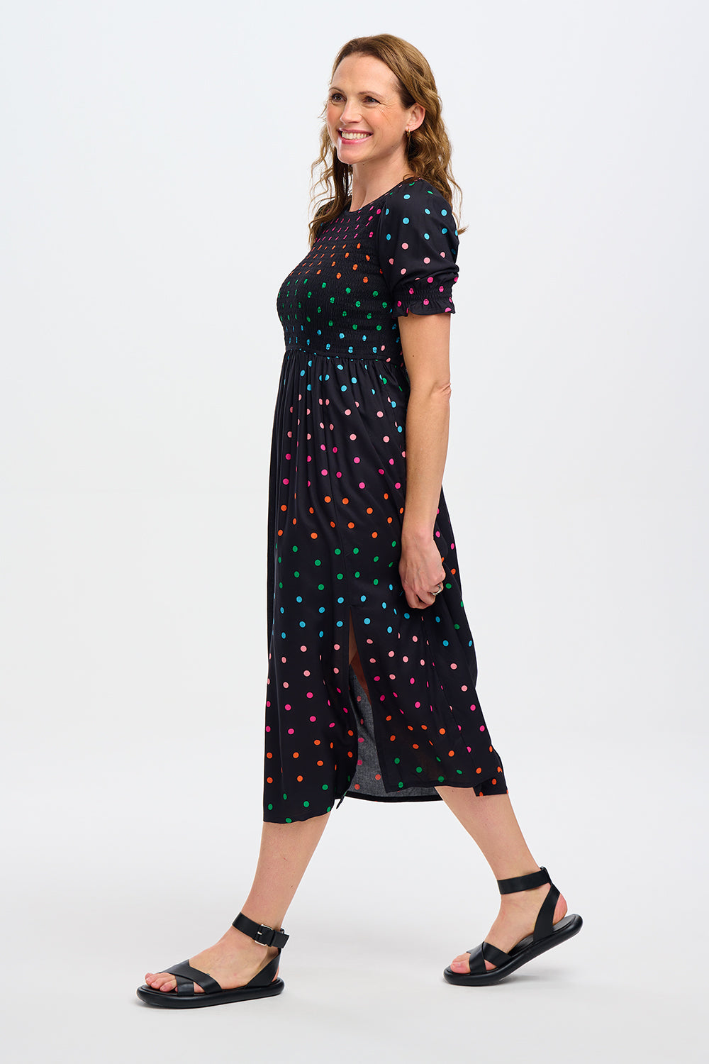 A model wearing a midi shirred dress with a black and rainbow polka print. The dress features a shirred bodice, elasticated puff sleeves, and a split hem.
