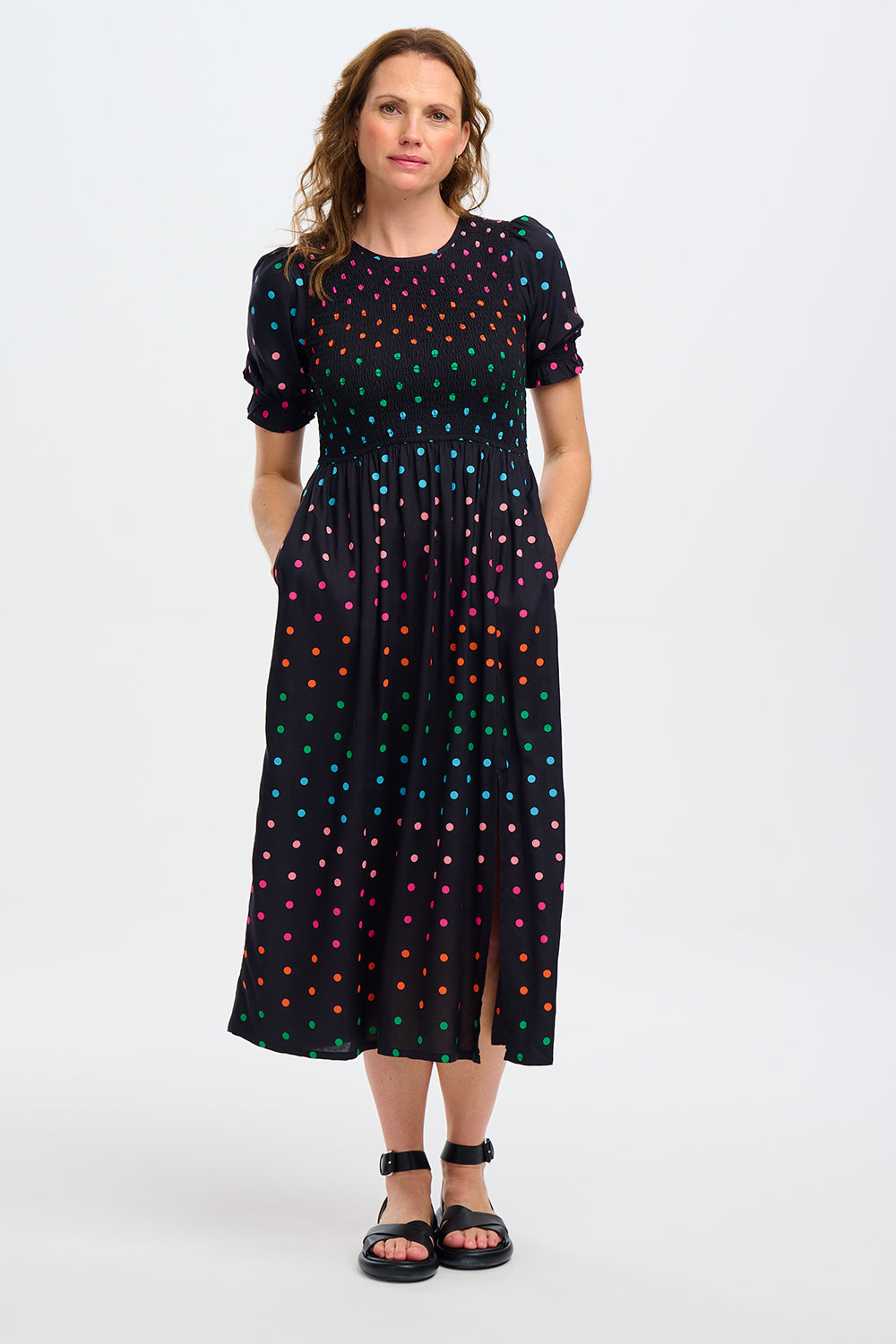 A model wearing a midi shirred dress with a black and rainbow polka print. The dress features a shirred bodice, elasticated puff sleeves, and a split hem.