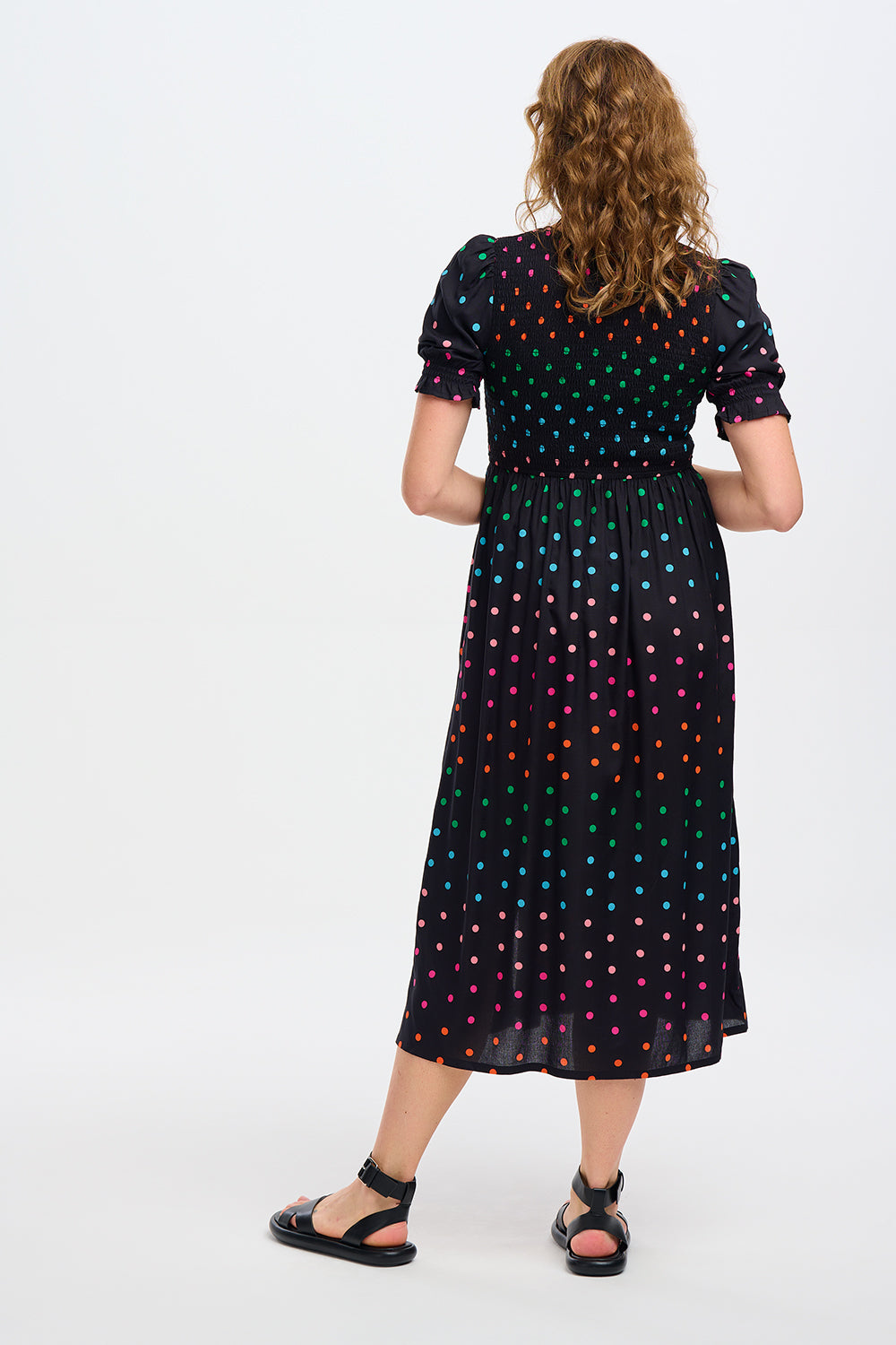 A model wearing a midi shirred dress with a black and rainbow polka print. The dress features a shirred bodice, elasticated puff sleeves, and a split hem.